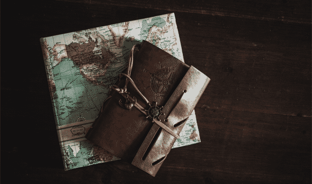 A leather-bound journal with a compass charm sits on top of a vintage map on a dark wooden surface.